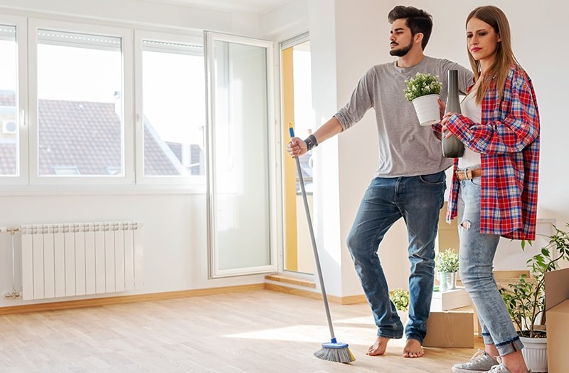 Young man with a broom and a woman with flower pot standing inside of a house