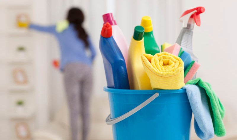 bucket full of chemical bottles and at back woman wiping a surface