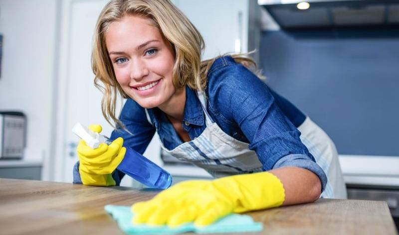 Woman in blue dress and yellow gloves holding bottle and cloth in her hand