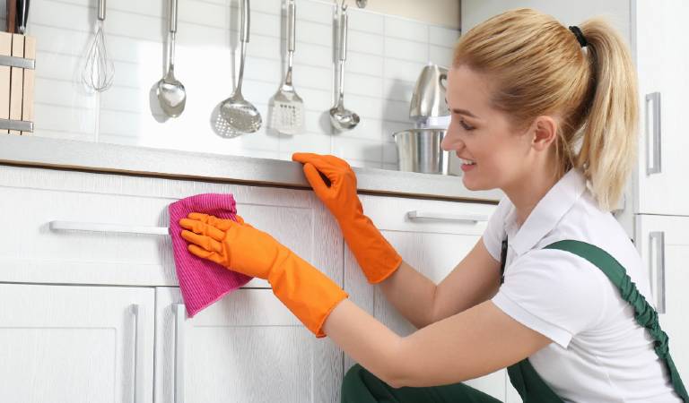 Woman in orange gloves scrubbing kitchen cabinet with red cloth
