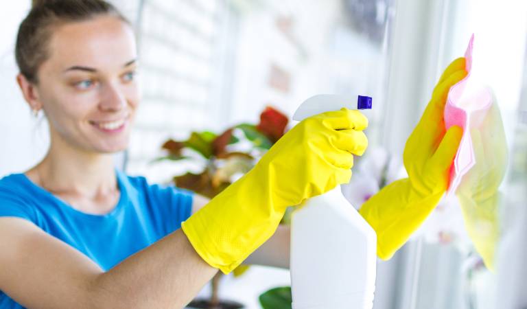 Woman in blue top holding white bottle in her hand scrubbing mirror with cloth