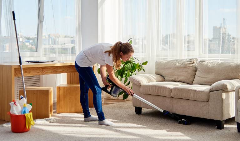 Woman in white t-shirt and blue jeans vacuuming below her sofa