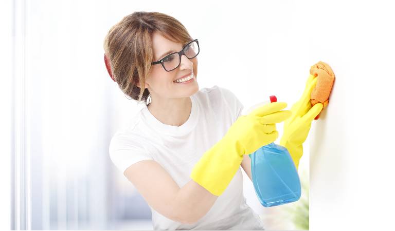 Woman in white top and yellow gloves holding a blue bottle and orange scrubber in her hand.