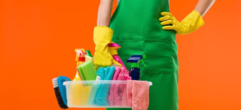 Woman in green uniform holding a yellow basket filled with tools and bottles.