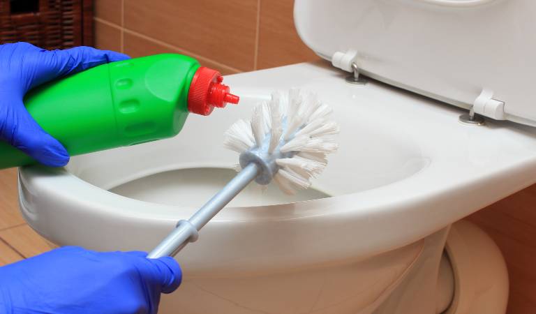 Hand in blue gloves holding a green bottle with red cap and brush cleaning toilet bowl.