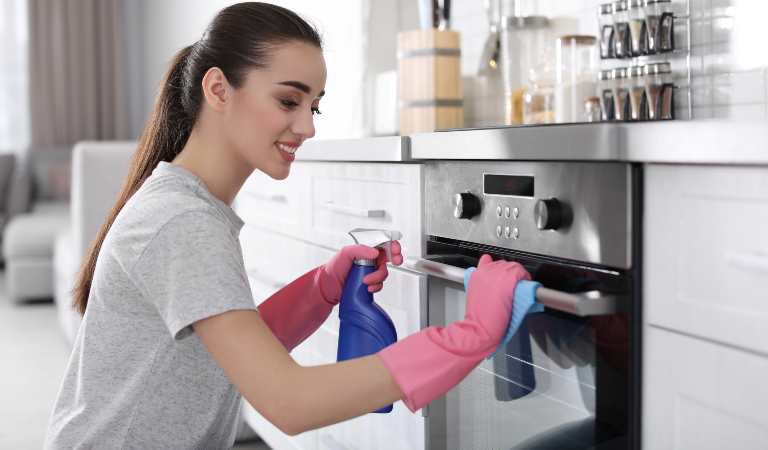 Woman in pink glove holding a blue bottle cleaning oven inside her kitchen