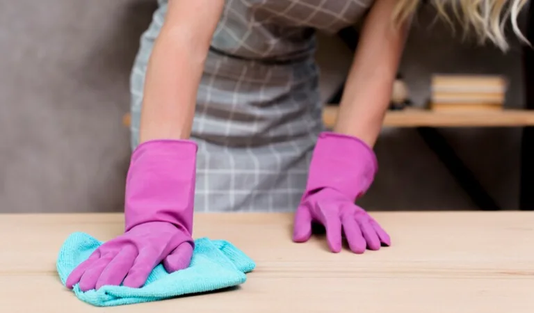 Lady in purple gloves cleaning wooden table with blue cloth.