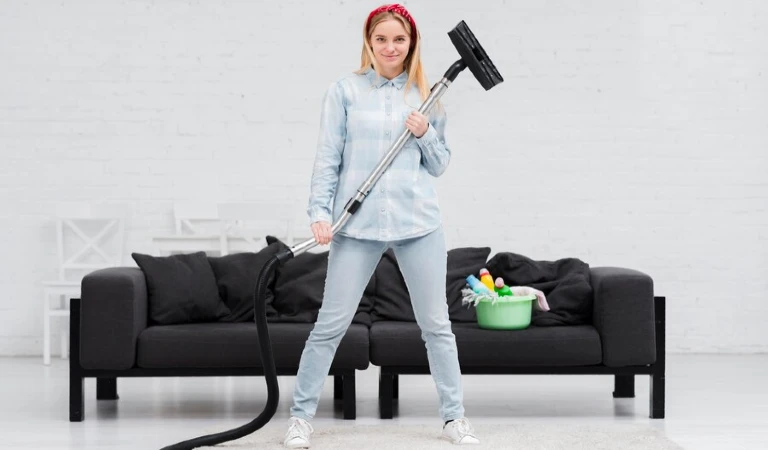 A lady holding a vacuum cleaner in her hand in front of a sofa.