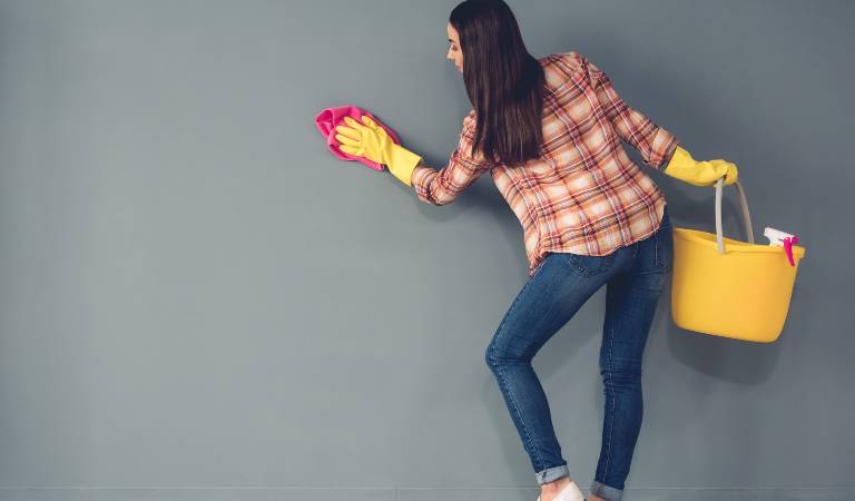 Woman in check shirt and blue jeans holding a yellow basket scrubbing wall.