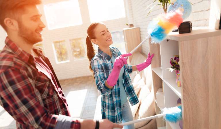 Man and a woman both in check shirt dusting wooden cabinets with a duster.