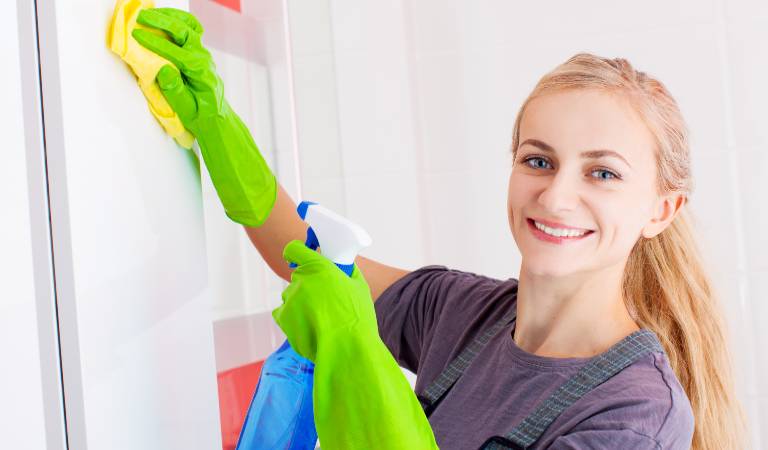 Smiling Woman in green gloves holding a blue bottle and yellow cloth in her hand.