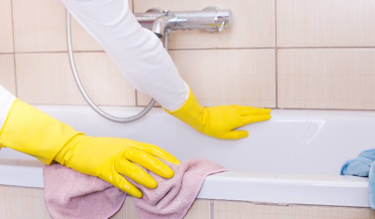 Woman in white top and yellow gloves cleaning bathtub with a light purple cloth.