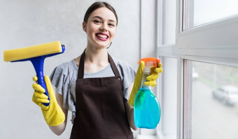 Young smiling lady in yellow gloves holding a bottle and a brush in her hand.