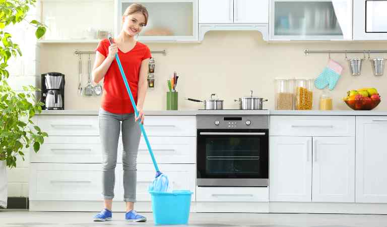 Young woman in red top and jeans holding a stick and blue basket on the floor insider her kitchen