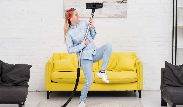 Woman in shirt and jeans playing with mop behind yellow sofa.