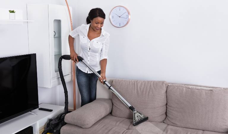Woman in white top and blue jeans cleaning couch with vacuum cleaner.