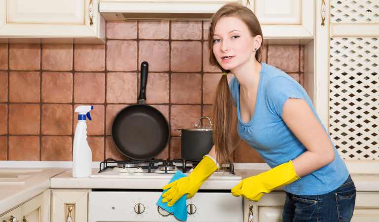 Woman in blue top and jeans cleaning kitchen cabinet with a cloth.