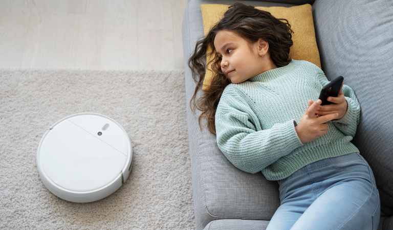 Small girl on sofa using her mobile and looking to a robot vacuum cleaner.