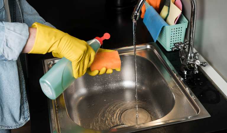 Woman in yellow glove holding a bottle cleaning kitchen sink
